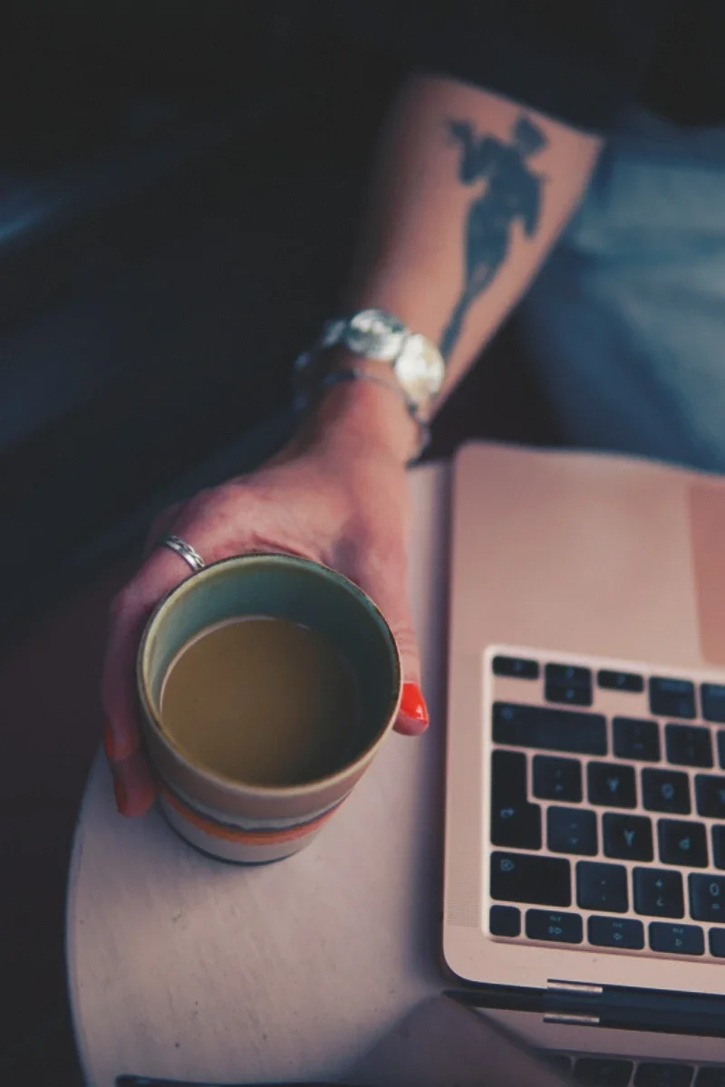 Hand med kaffekopp bredvid en laptop i en varm arbetsmiljö.